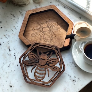 May include: Two wooden coasters with a bee design. The coasters are hexagonal and have a dark brown wood finish. The bee design is laser cut into the wood and is a light brown color.