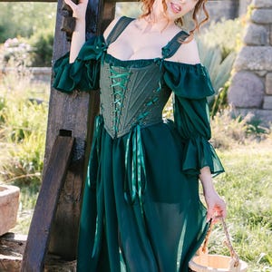 May include: A dark green medieval-style dress with a corset bodice and flowing skirt. The dress features off-the-shoulder sleeves and ribbon details. A small wooden bucket is held in the hand. The background includes a wooden structure and stone buildings.
