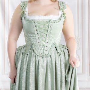 Puede incluir: Un vestido hist&oacute;rico verde claro con corpi&ntilde;o de cors&eacute; y falda amplia. El vestido presenta una parte delantera con cordones, ribete con volantes y un estampado floral. El vestido largo tiene una falda en capas con volantes en el dobladillo. La modelo lleva zapatos marrones.