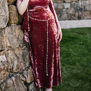 May include: A burgundy velvet dress with short sleeves and a flowing design. The dress features a gold-colored headband and a delicate pearl belt. The model is leaning against a stone wall, creating a romantic and elegant aesthetic.