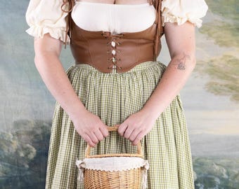 Könnte beinhalten: Ein braunes Lederkorsett mit Schnürung wird mit einer weißen Bluse und einem grün-weiß karierten Rock kombiniert. Ein kleiner Weidenkorb mit weißer Spitzentrimmung vervollständigt das Outfit.
