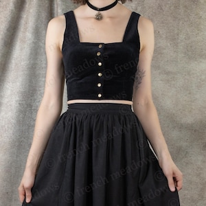 May include: A black two-piece outfit featuring a velvet crop top with gold buttons and a square neckline, paired with a flowing midi skirt. The outfit is completed with black boots. The background is a neutral, textured fabric.