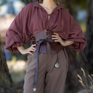 May include: A burgundy peasant blouse with puffed sleeves, paired with brown linen pants and leather boots. A brown leather corset cinches the waist, and a long belt hangs down. A pocket watch hangs from the belt. The outfit is suitable for Renaissance fairs or cosplay.