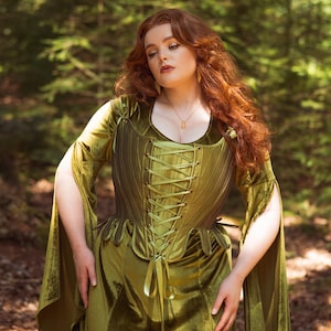 May include: An olive green velvet dress featuring a structured corset-style bodice with front lace-up detailing, a scoop neckline, and long, split bell sleeves. The garment has a fitted waist and a floor-length skirt with a soft, light-reflecting finish.