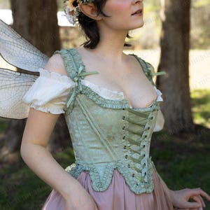 May include: A green corset with a lace-up front and a pink skirt. The corset has a ruffled trim and a bow on the shoulder straps. The skirt is a simple, flowing style.