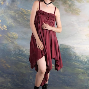 May include: A burgundy-colored dress with spaghetti straps and a gathered bodice. The dress has an asymmetrical hemline and is paired with black lace-up boots. The background features a landscape painting.