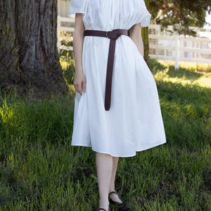 Puede incluir: Un vestido blanco con hombros descubiertos y cinturón marrón. El vestido tiene mangas cortas y una falda fluida. El cinturón se ciñe a la cintura con una hebilla circular. La persona lleva sandalias negras.