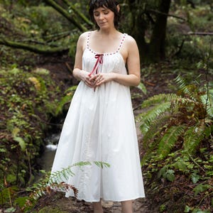 Puede incluir: Vestido blanco de algodón con tirantes finos, adornado con lunares rojos en el escote y un lazo de cinta roja. El vestido llega a la rodilla y tiene una falda fluida. La imagen está ambientada en un entorno natural al aire libre.