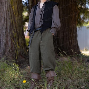 May include: A person wearing a historical costume. The outfit includes a dark vest, a long-sleeved shirt, olive green pants, and brown boots. The person is standing in front of large trees.