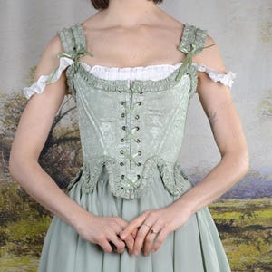 Puede incluir: Un cors&eacute; verde claro con un estampado floral, ribetes de encaje y lazos de cinta. El cors&eacute; se combina con una falda a juego y una blusa blanca con volantes. El conjunto tiene una est&eacute;tica vintage o hist&oacute;rica.