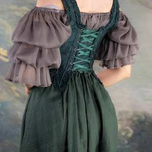 May include: A dark green corset-style bodice with green lacing, paired with a matching dark green skirt. The ensemble includes gray, layered, off-the-shoulder sleeves. The outfit is reminiscent of historical or fantasy attire.