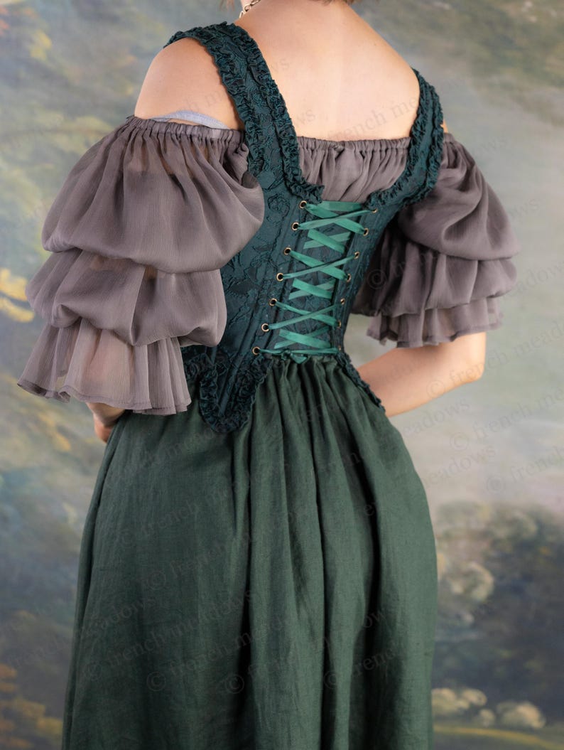 May include: A dark green corset-style bodice with green lacing, paired with a matching dark green skirt. The ensemble includes gray, layered, off-the-shoulder sleeves. The outfit is reminiscent of historical or fantasy attire.
