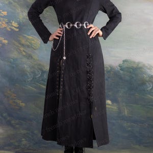 May include: A long black dress with a V-neck and long sleeves. The dress features a silver chain belt and a slit up the front. The dress has a lace-up detail on the side and is worn with black boots. The background is a painted landscape.