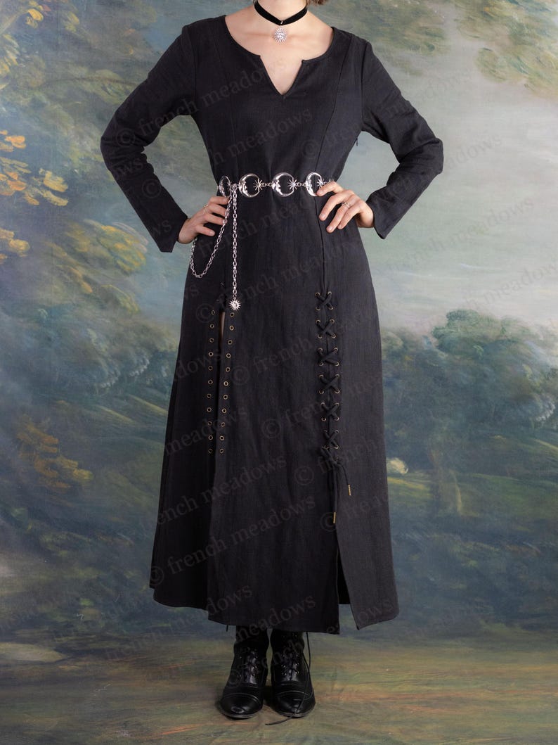 May include: A long black dress with a V-neck and long sleeves. The dress features a silver chain belt and a slit up the front. The dress has a lace-up detail on the side and is worn with black boots. The background is a painted landscape.