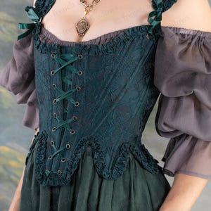 May include: A dark teal corset with a floral pattern, lace-up front, and ruffled edges. The corset has dark green ribbon accents and is paired with gray, off-the-shoulder sleeves and a dark green skirt. A gold-toned necklace with a pendant is also visible.