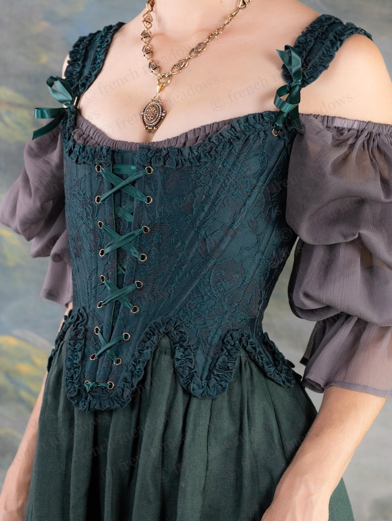 May include: A dark teal corset with a floral pattern, lace-up front, and ruffled edges. The corset has dark green ribbon accents and is paired with gray, off-the-shoulder sleeves and a dark green skirt. A gold-toned necklace with a pendant is also visible.