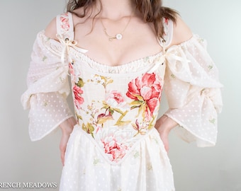 May include: A white off-the-shoulder dress with a floral print bodice and a sheer overlay. The dress has a fitted bodice and a flowing skirt.