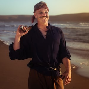 Peut inclure: Une personne souriante, vêtue d'un costume de pirate sur une plage. Ils portent une chemise bleu foncé, un pantalon marron et un bandana rouge. Une ceinture noire et un pistolet jouet complètent le costume. L'arrière-plan montre la mer et un coucher de soleil.