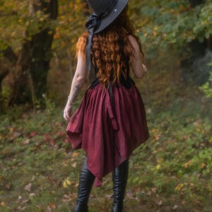 May include: A black witch hat with a wide brim and a black ribbon, paired with a burgundy skirt and black boots. The outfit is worn in a forest setting, evoking a mystical or fantasy theme.