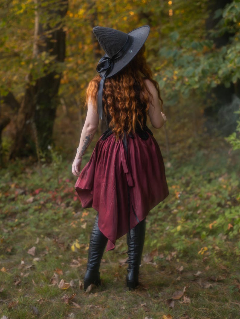 May include: A black witch hat with a wide brim and a black ribbon, paired with a burgundy skirt and black boots. The outfit is worn in a forest setting, evoking a mystical or fantasy theme.