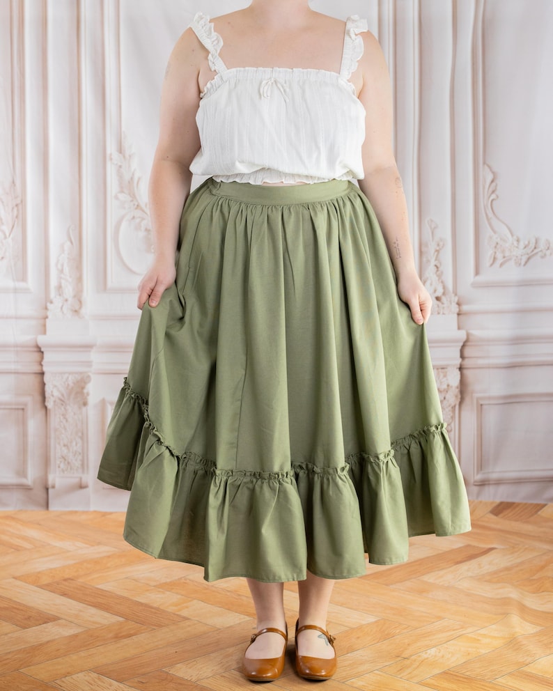 May include: A white, ruffled crop top with spaghetti straps paired with a long, olive green skirt with a ruffled hem. The outfit is completed with brown Mary Jane shoes. The clothing is likely suitable for a casual or vintage-inspired aesthetic.