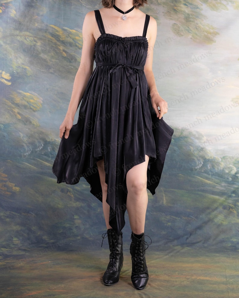 Puede incluir: Vestido negro con dobladillo asim&eacute;trico. El vestido tiene un corpi&ntilde;o fruncido, tirantes finos y un cintur&oacute;n anudado. La persona lleva botas negras con cordones y un collar gargantilla negro.