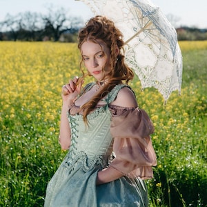 Puede incluir: Una mujer con un vestido verde claro y marr&oacute;n con un parasol de encaje est&aacute; en un campo de flores amarillas. El vestido tiene un corpi&ntilde;o estilo cors&eacute; y mangas con volantes. El parasol es blanco con detalles de encaje.