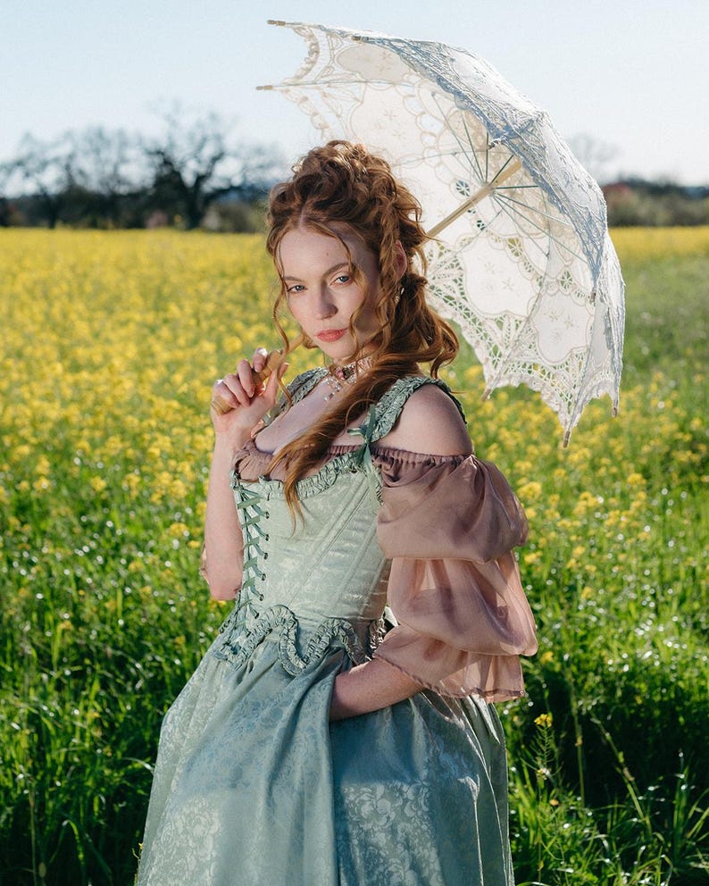 Puede incluir: Una mujer con un vestido verde claro y marr&oacute;n con un parasol de encaje est&aacute; en un campo de flores amarillas. El vestido tiene un corpi&ntilde;o estilo cors&eacute; y mangas con volantes. El parasol es blanco con detalles de encaje.