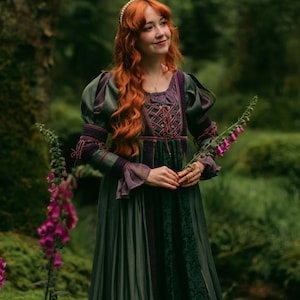 May include: A floor-length dress in shades of dark green and purple, featuring a lace-up bodice with crisscross detailing, long sleeves with ruffled cuffs, and a textured central panel. The garment is worn with a pearl-embellished headband.