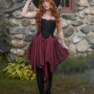 May include: A black corset top with lace-up detailing and a burgundy skirt. The outfit is paired with a black hat and knee-high black boots. The woman is standing in front of a stone building.