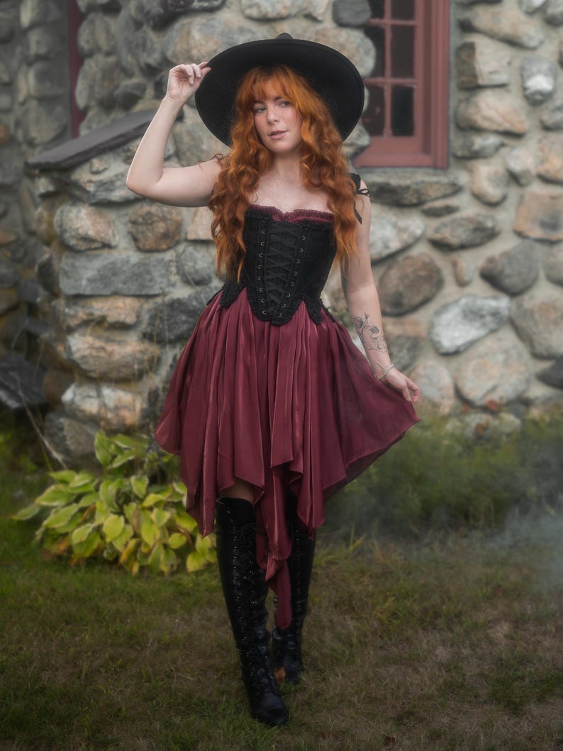May include: A black corset top with lace-up detailing and a burgundy skirt. The outfit is paired with a black hat and knee-high black boots. The woman is standing in front of a stone building.