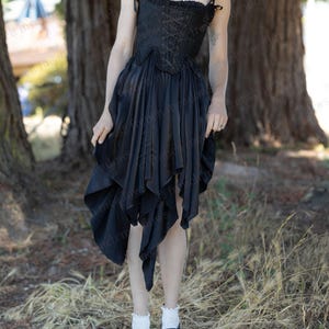 Puede incluir: Un vestido negro con un corpi&ntilde;o estilo cors&eacute; y una falda asim&eacute;trica fluida. El vestido presenta un frente con cordones y tirantes delicados. La modelo lleva calcetines blancos y zapatos negros con detalles blancos. El fondo incluye &aacute;rboles y hierba seca.