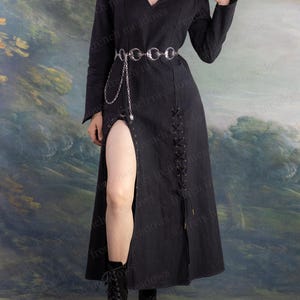 May include: A long black dress with a high slit, long sleeves, and a V-neck. The dress features a silver chain belt and is paired with black boots. The background is a painted landscape.