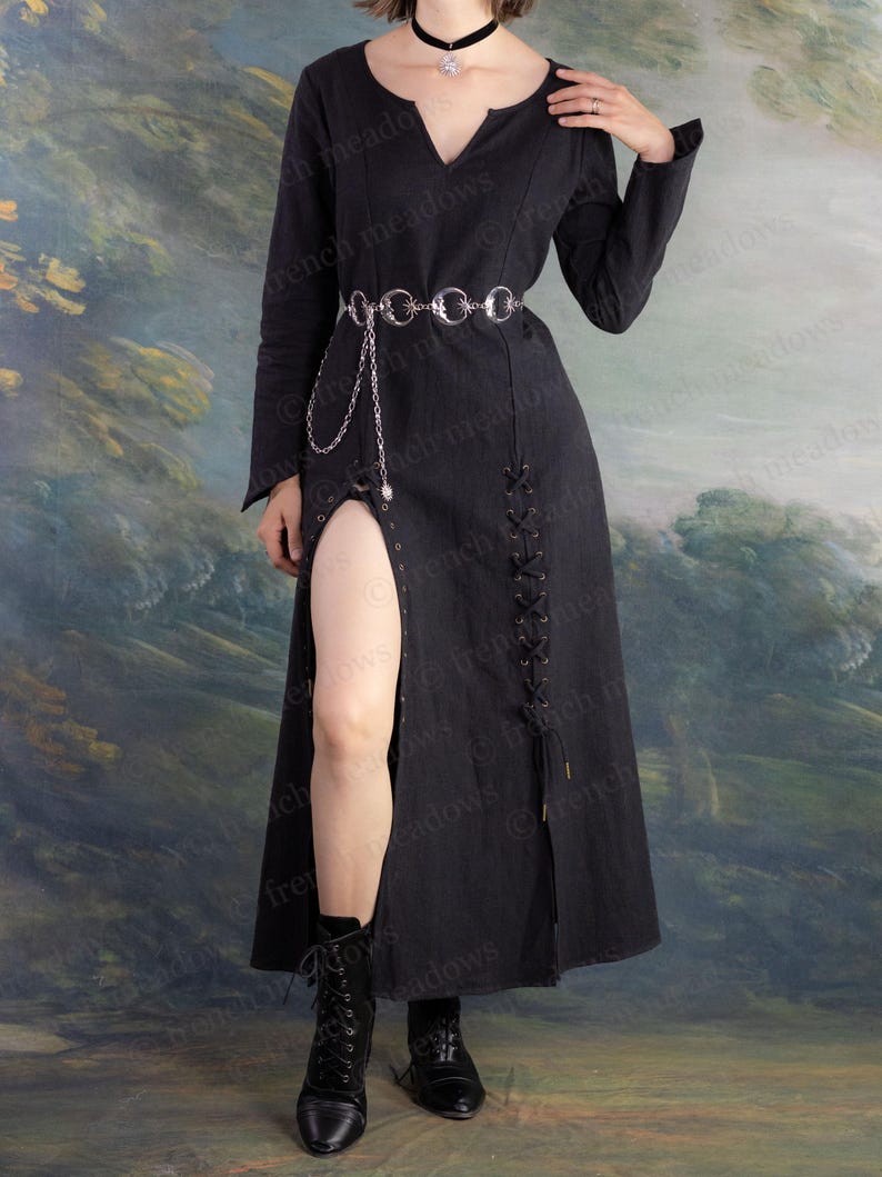May include: A long black dress with a high slit, long sleeves, and a V-neck. The dress features a silver chain belt and is paired with black boots. The background is a painted landscape.