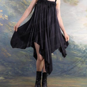 Puede incluir: Vestido negro asim&eacute;trico con tirantes finos. El vestido tiene un dobladillo fluido en forma de pa&ntilde;uelo y se combina con botas negras con cordones. El fondo es un paisaje pintado con colores apagados.