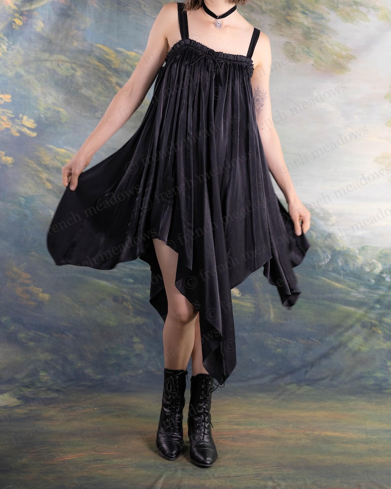 Puede incluir: Vestido negro asim&eacute;trico con tirantes finos. El vestido tiene un dobladillo fluido en forma de pa&ntilde;uelo y se combina con botas negras con cordones. El fondo es un paisaje pintado con colores apagados.