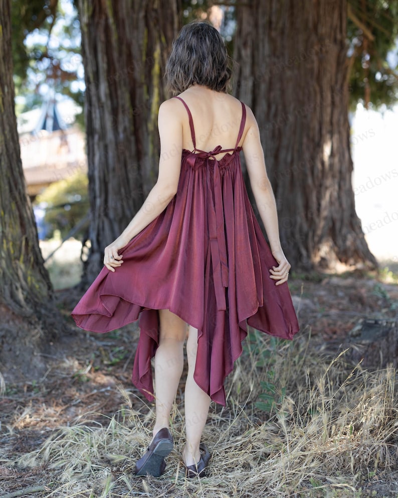 May include: A burgundy-colored dress with spaghetti straps and a handkerchief hem. The dress features a bow detail at the back and is made of a flowing fabric. The model is wearing dark shoes and is standing in a wooded area.