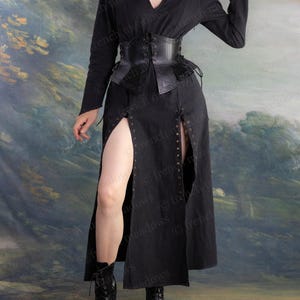 May include: A black medieval-style outfit featuring a long-sleeved dress with a V-neck, a corset, and a long skirt with high slits. The outfit is paired with black boots. The corset is made of leather and has intricate detailing.