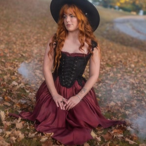 May include: A person wearing a black corset with a burgundy skirt and a black hat, sitting on a bed of fallen leaves. The corset has a lace-up front and the skirt is flowing. The hat is wide-brimmed.