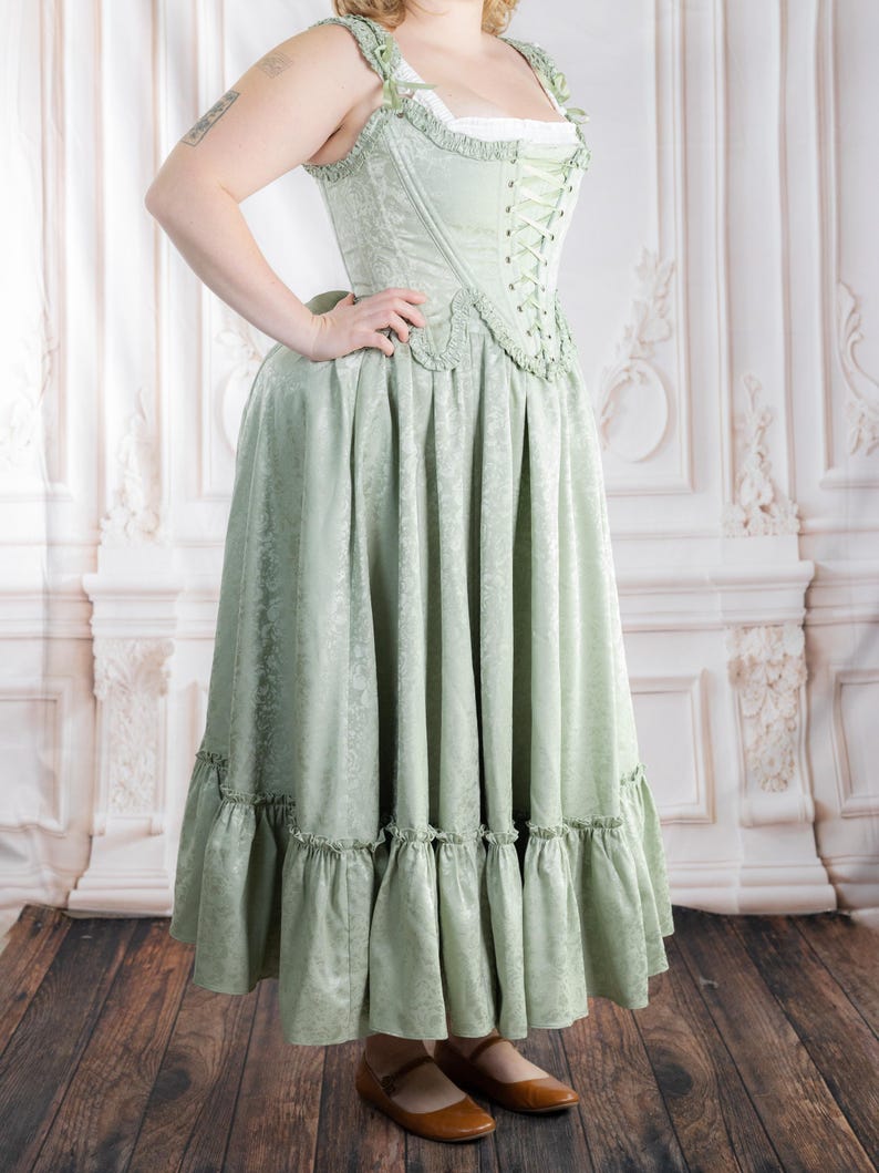 Puede incluir: Un vestido cors&eacute; verde claro con un estampado floral. El vestido presenta un corpi&ntilde;o con cordones, una falda amplia con volantes y una camiseta interior blanca. El vestido se combina con bailarinas marrones. El vestido es aproximadamente hasta la rodilla.