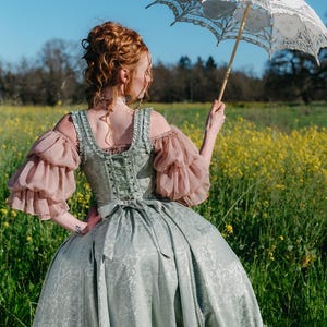 Puede incluir: Un vestido hist&oacute;rico verde claro y rosa con un parasol de encaje blanco. El vestido presenta un corpi&ntilde;o con cors&eacute;, mangas abullonadas y una falda amplia. El parasol es blanco con intrincados detalles de encaje. La escena se desarrolla en un campo de flores amarillas.