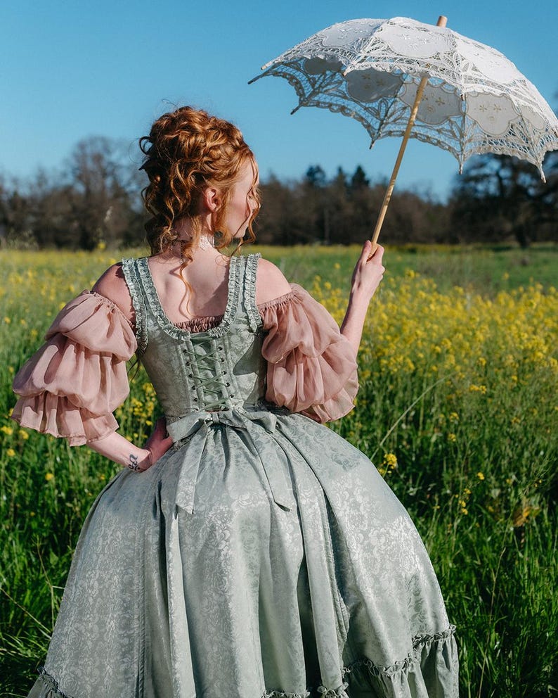 Puede incluir: Un vestido hist&oacute;rico verde claro y rosa con un parasol de encaje blanco. El vestido presenta un corpi&ntilde;o con cors&eacute;, mangas abullonadas y una falda amplia. El parasol es blanco con intrincados detalles de encaje. La escena se desarrolla en un campo de flores amarillas.