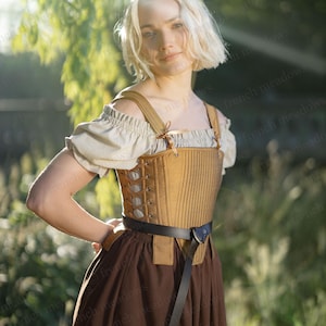 Puede incluir: Un corsé color beige con correas y cordones marrones, usado sobre una blusa crema y una falda marrón. Un cinturón negro ciñe la cintura. El atuendo evoca épocas históricas o fantásticas.