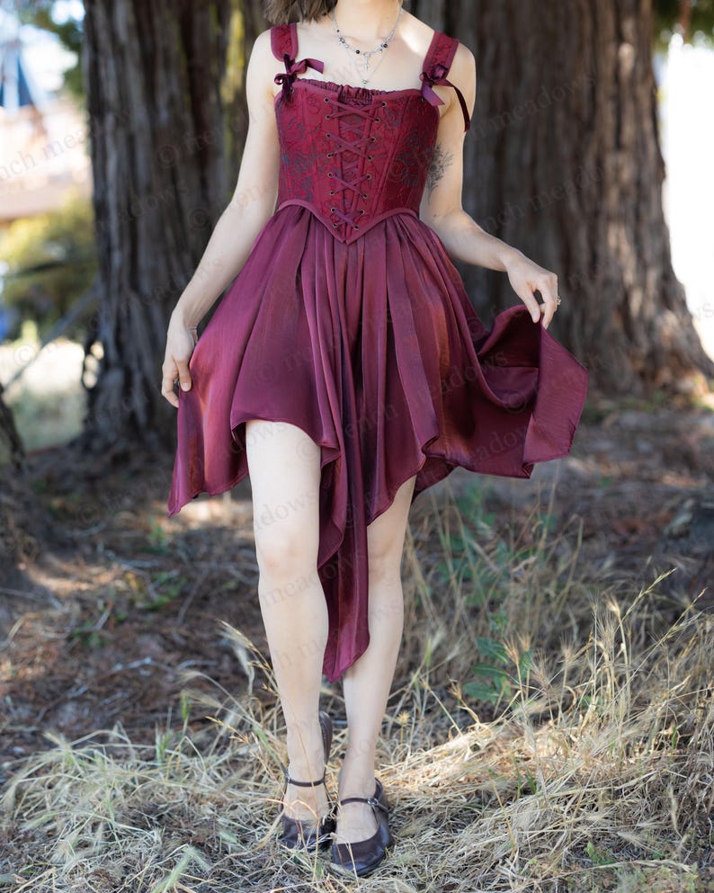 May include: A burgundy dress with a corset-style bodice and a flowing, asymmetrical skirt. The dress features delicate ribbon straps and a lace-up front. The model is wearing dark brown Mary Jane shoes. The dress is suitable for special occasions.