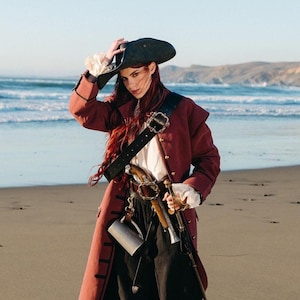 Puede incluir: Un abrigo de pirata granate con detalles negros, usado con un sombrero y pantalones negros. El atuendo incluye un cinturón de cuero negro con hebilla plateada, una pistola y una espada. La escena se desarrolla en una playa de arena con el océano al fondo.