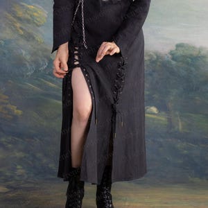 May include: A black dress with long sleeves and a high slit, featuring lace-up detailing on the sides. A silver chain belt with circular links and a pair of black lace-up boots complete the outfit. The dress is made of a dark fabric.