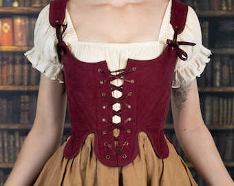 May include: A deep burgundy corduroy bodice featuring a front lace-up closure with metal grommets and ribbon ties. The garment has wide shoulder straps secured with small ribbon bows and is worn over a cream-colored short-sleeved blouse and a tan skirt.