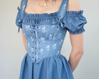 Puede incluir: Top y vestido de corsé azul claro con estampado floral blanco. El corsé presenta un frente con cordones y se combina con mangas con hombros descubiertos y una falda a juego. Las correas están adornadas con pequeños lazos.