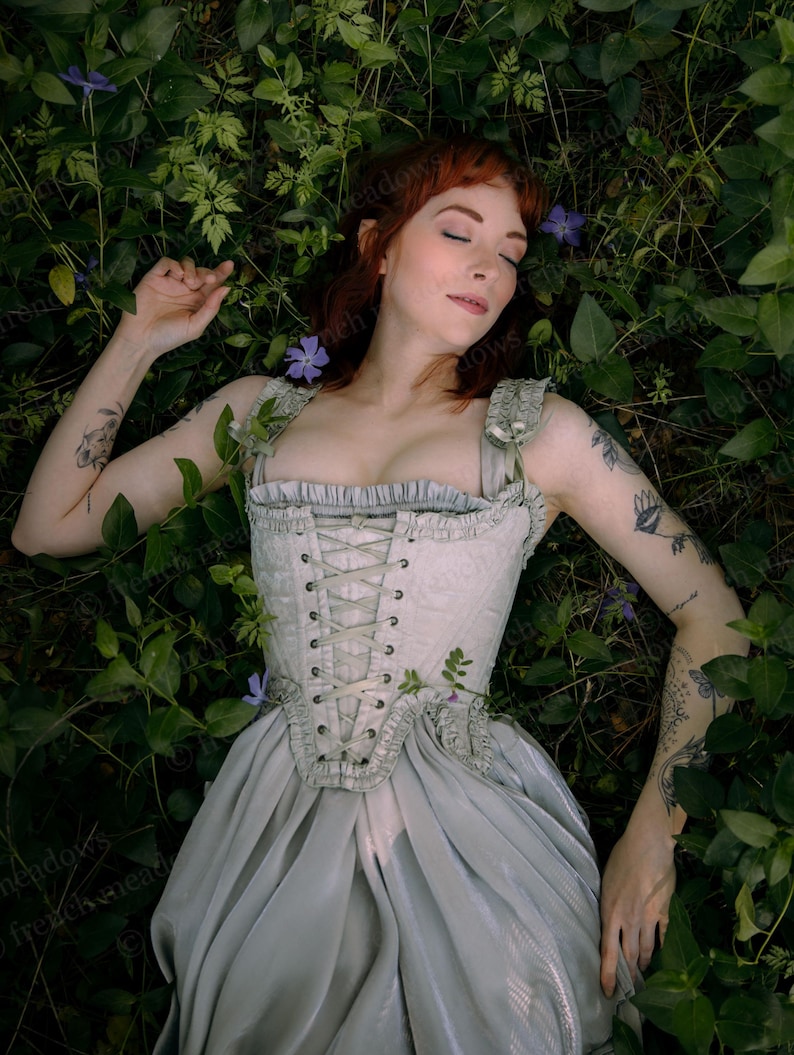 Puede incluir: Un top de estilo cors&eacute; verde claro y una falda a juego, entre hojas verdes y peque&ntilde;as flores moradas. El cors&eacute; tiene un frente con cordones y detalles decorativos. El conjunto es suave y rom&aacute;ntico.