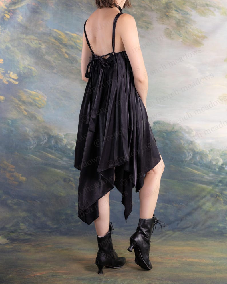 Puede incluir: Vestido negro asim&eacute;trico con tirantes finos y un lazo en la espalda. El vestido se combina con botines negros con un tac&oacute;n peque&ntilde;o. El fondo presenta un paisaje suave y pict&oacute;rico.